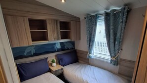 Ein kleiner Raum mit zwei Betten, einem kleinen Nachttisch mit Pflanze und einem Fenster mit Vorhängen.