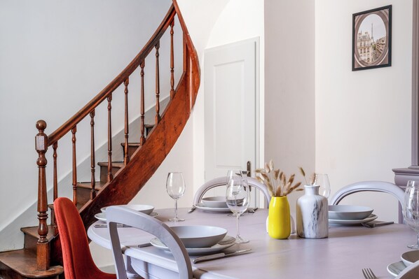 A dining area with a wooden staircase, white chairs, a table set for two, and a vase with dried pampas grass.