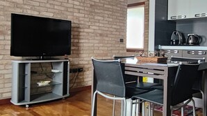 Ein geräumiges Wohnzimmer mit Backsteinwand, Holzbalken, einem Flachbildfernseher, einem Esstisch mit Stühlen und einer Küchenzeile mit Schränken und Geräten.