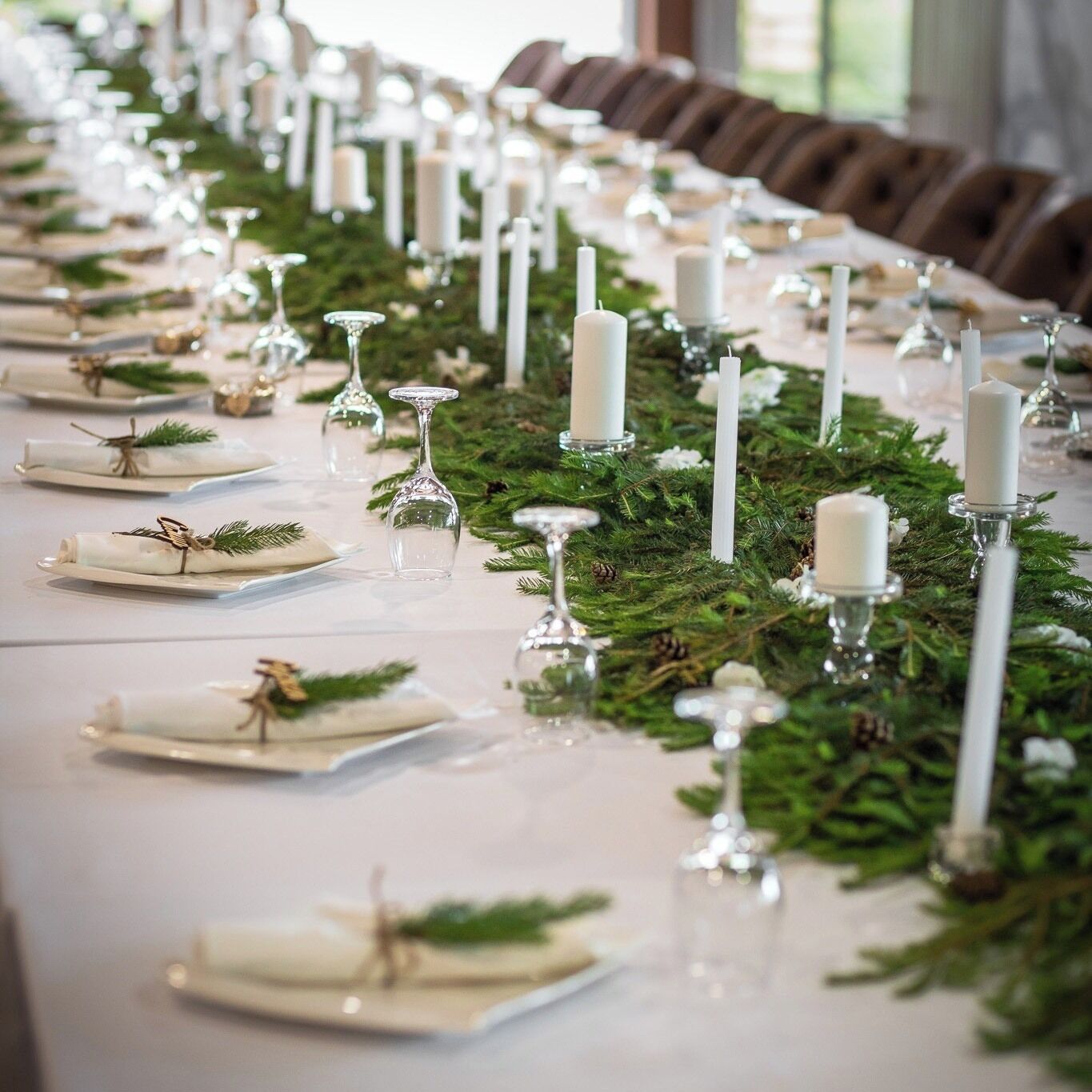 A long dining table set with white plates, clear glassware, and white candles, adorned with greenery and pine cones.