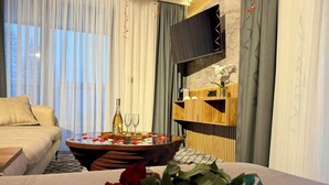 Une chambre d’hôtel moderne avec un lit, une table ronde décorée de pétales de rose, une bouteille de vin et des verres.