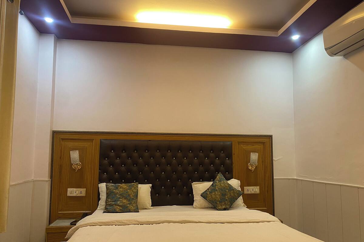 A hotel room with a large bed, a wooden headboard, two bedside tables, and a wall-mounted air conditioning unit.
