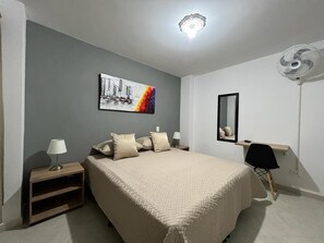 A bedroom with a bed, bedside tables, a desk, a chair, a mirror, and a ceiling fan. - AMOBLADO LOS MOLINOS (Medellín)