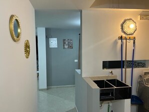 A room with a white wall, a black cabinet, and a blue chair. There is a framed picture on the wall and a light fixture above the cabinet. - AMOBLADO LOS MOLINOS (Medellín)