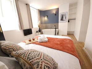 Habitación de hotel con una cama grande, una mesita de noche con una taza y una toalla, un baño con bañera y ducha, y un televisor montado en la pared.