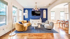 Un salon moderne avec un mur d’accent bleu, un téléviseur à écran plat, un canapé beige, un fauteuil gris et un coin repas avec des chaises en bois.