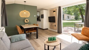 A modern living room with a sofa, a dining table, and a TV. There is a kitchen area with cabinets and a TV. The room has a large window with curtains. - Chalet With a Dishwasher, in a Car-free Park (Vlierden)