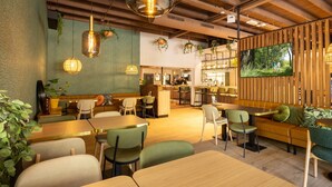 A restaurant with wooden tables and chairs, hanging pendant lights, and a large TV screen on the wall. - Chalet With a Dishwasher, in a Car-free Park (Vlierden)