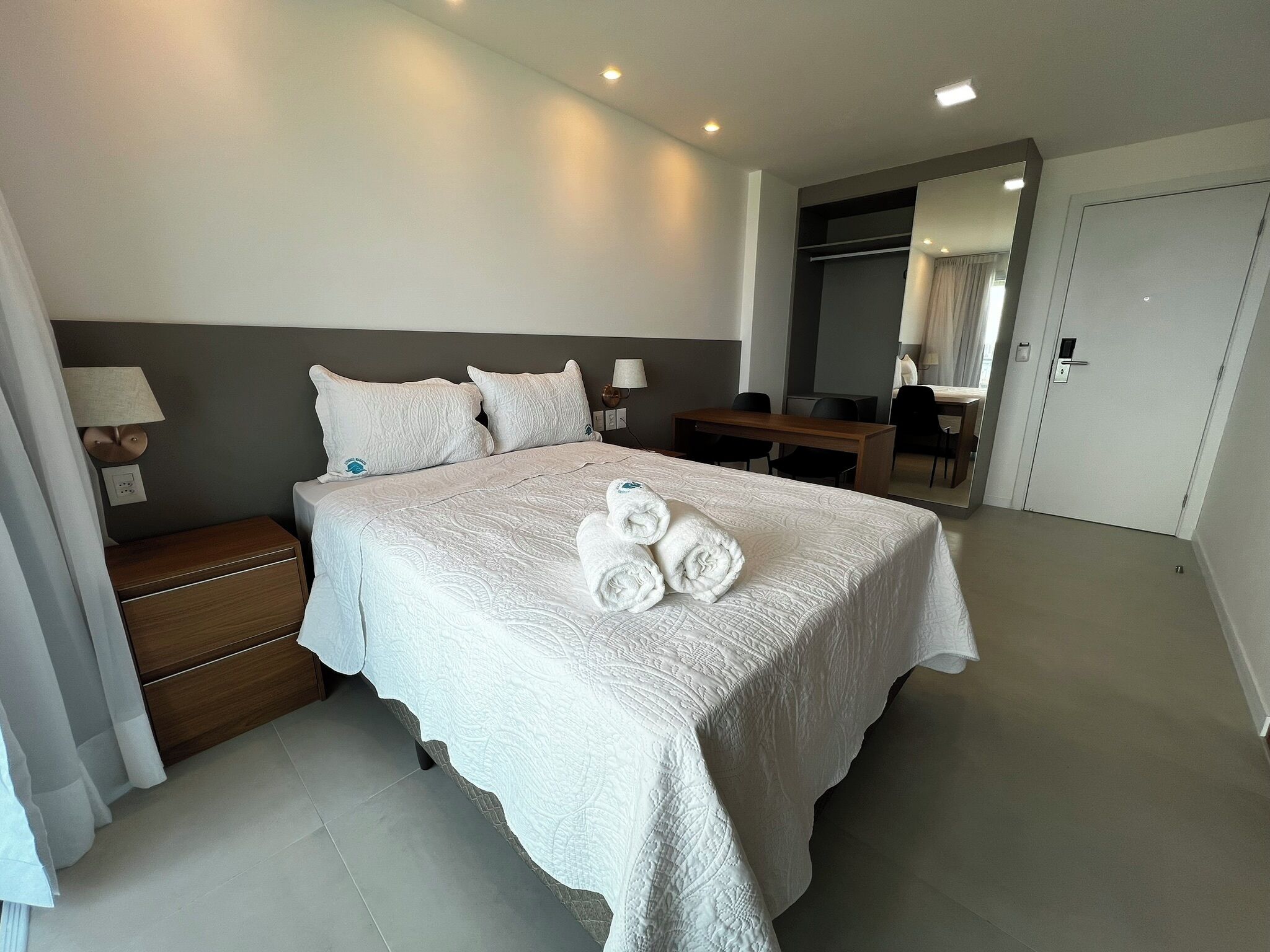 A modern hotel room with a large bed, bedside lamps, a wooden nightstand, and a desk with chairs.