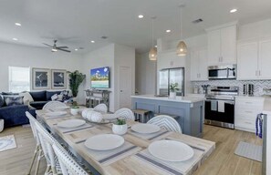 Une cuisine moderne avec des meubles blancs, un îlot central et un coin repas agrémenté d’une longue table dressée pour six personnes.