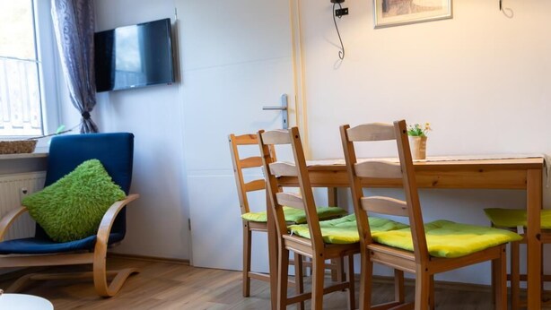 A room with a wooden dining table and four chairs, a blue chair with a green cushion, a television mounted on the wall, and a window with curtains.