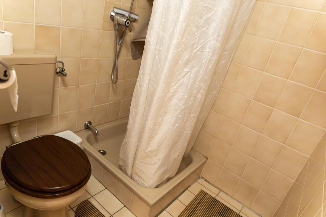 A bathroom with a wooden toilet, a shower with a white shower curtain, and beige tiled walls.