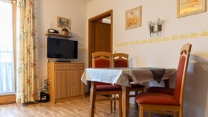 Un comedor con una mesa dispuesta para dos personas, sillas de madera, un televisor sobre un mueble y una ventana con cortinas.