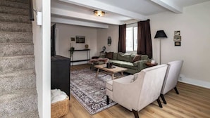 A spacious living area with a staircase, a green sofa, a white armchair, a coffee table, and a floor lamp. - 3BR Getaway, Sleeps 6 | 2-3 Miles to ISU (Ames)