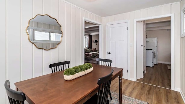 A dining area with a wooden table, chairs, and a decorative mirror. There are white paneled walls and a ceiling light fixture. - 3BR Getaway, Sleeps 6 | 2-3 Miles to ISU (Ames)
