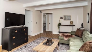 A spacious living room with a large flat-screen TV, a black dresser with gold knobs, a green sofa, and a wooden coffee table. - 3BR Getaway, Sleeps 6 | 2-3 Miles to ISU (Ames)