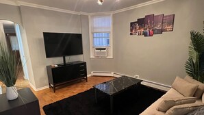 Una sala de estar con un televisor de pantalla plana, una mesa de centro negra, un sofá beige, una alfombra negra y un accesorio de iluminación en el techo.