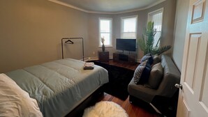 Un dormitorio con cama, televisor, sofá, planta y lámpara colgante.
