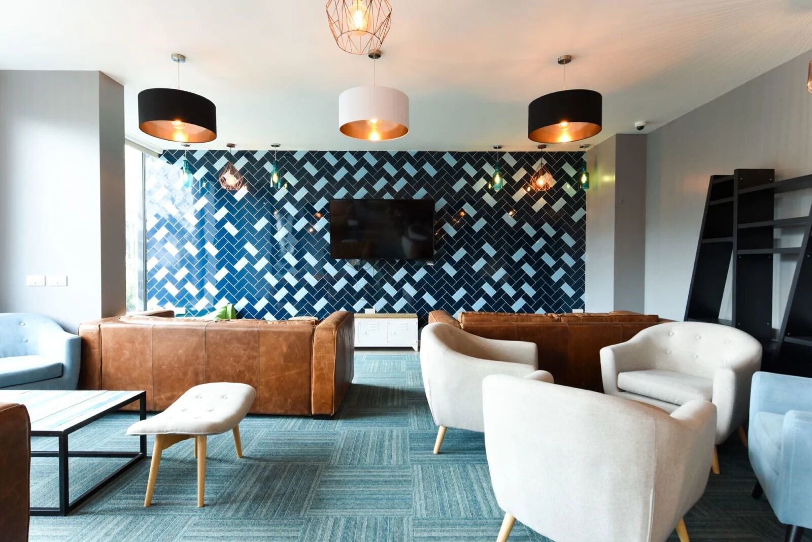 A modern lounge area with a herringbone patterned wall, a flat-screen TV, and contemporary furniture including a brown leather sofa, white armchairs, and a small table with a lamp.