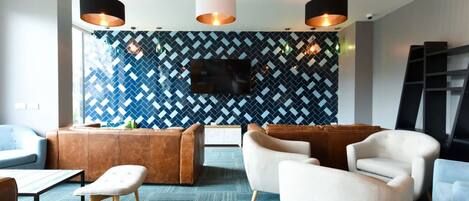A modern lounge area with a herringbone patterned wall, a flat-screen TV, and contemporary furniture including a brown leather sofa, white armchairs, and a small table with a lamp.