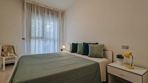 A bedroom with a bed, a bedside table with a lamp, a chair, and a window with curtains.