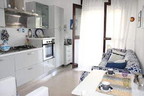 A modern kitchen with white cabinets, a stove, and an oven. There is a dining area with a table and chairs, and a sofa with patterned cushions.