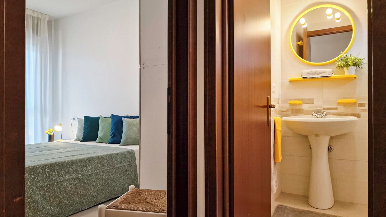 Ein Schlafzimmer mit einem Bett, Kissen und einem Nachttisch. Im Raum befindet sich ein Fenster mit Vorhängen.
