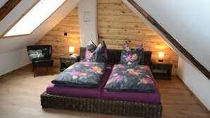 Un dormitorio tipo loft con techo inclinado, tragaluz, cama con ropa de cama floral, dos mesitas de noche con lámparas y una silla.