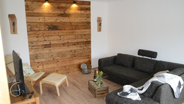 Una sala de estar con panel de pared de madera, un sofá gris, una mesa de centro de madera con un florero, un televisor sobre un mueble de madera y una silla.