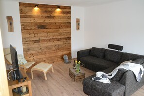 Een woonkamer met een houten wandpaneel, een grijze bank, een houten salontafel met een vaas, een televisie op een houten dressoir, en een stoel.