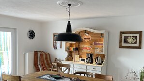 Un comedor con una mesa de madera, sillas, una lámpara colgante negra y una cocina al fondo con una cafetera y un letrero que dice "Superior Coffee".