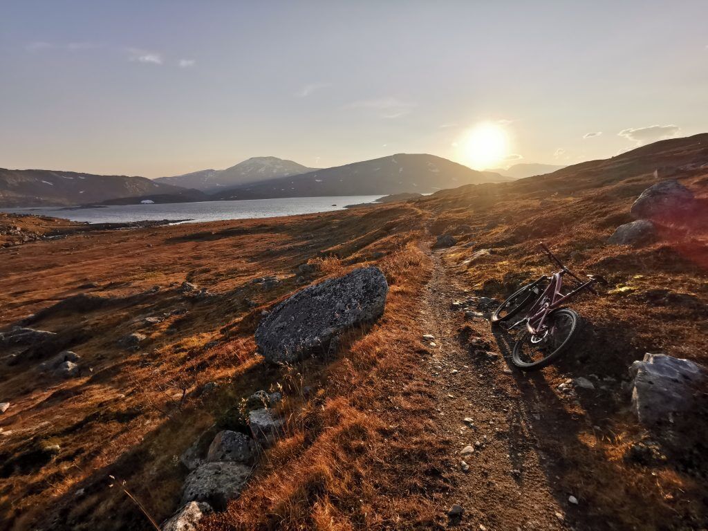 Uma trilha de montanha com uma bicicleta e uma rocha grande em primeiro plano, que leva a um lago e montanhas ao longe.