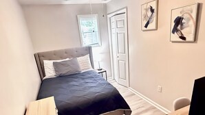 A bedroom with a bed, bedside table, chair, and a desk with a computer monitor. There are two paintings on the wall and a window with blinds.