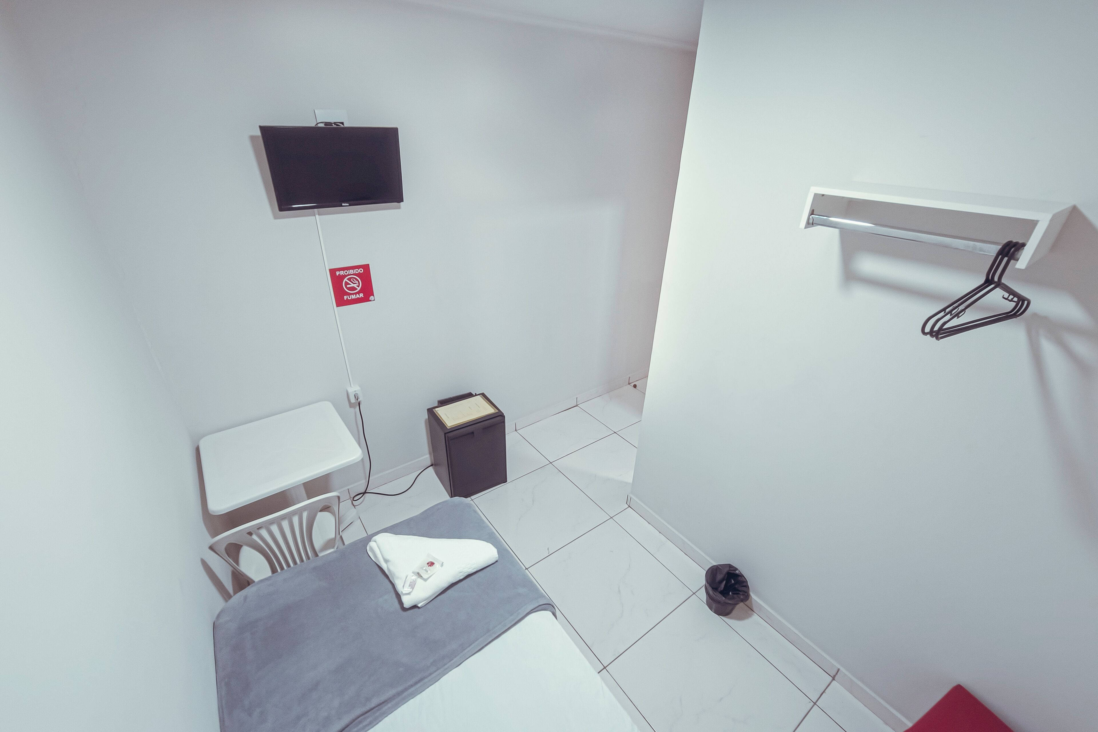 Ein kleines, sauberes Zimmer mit einem Bett, einem an der Wand befestigten Fernseher, einem kleinen Tisch und einem Mülleimer.