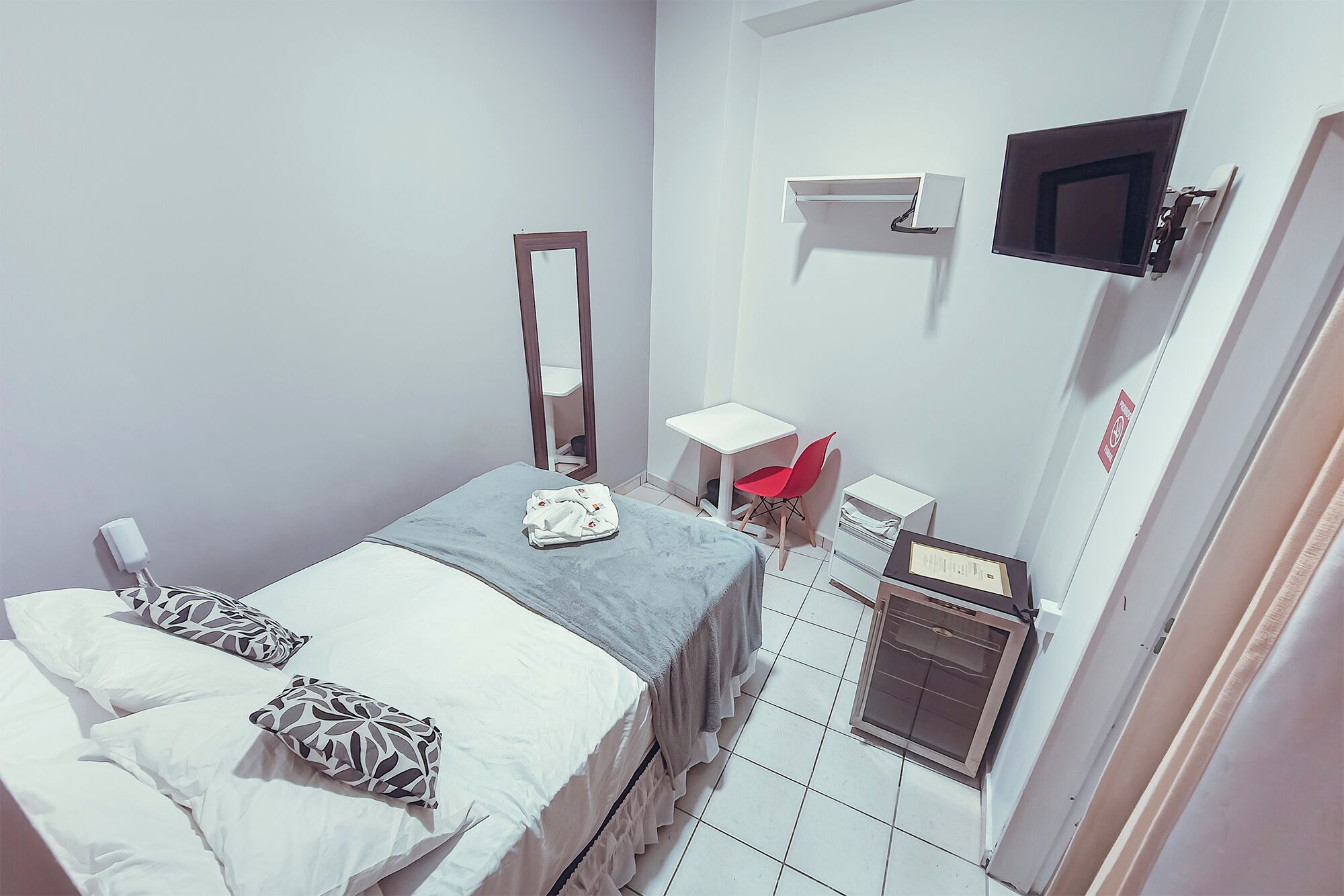 Un dormitorio pequeño y ordenado con cama, mesita de noche, silla, espejo, televisor y calefactor.