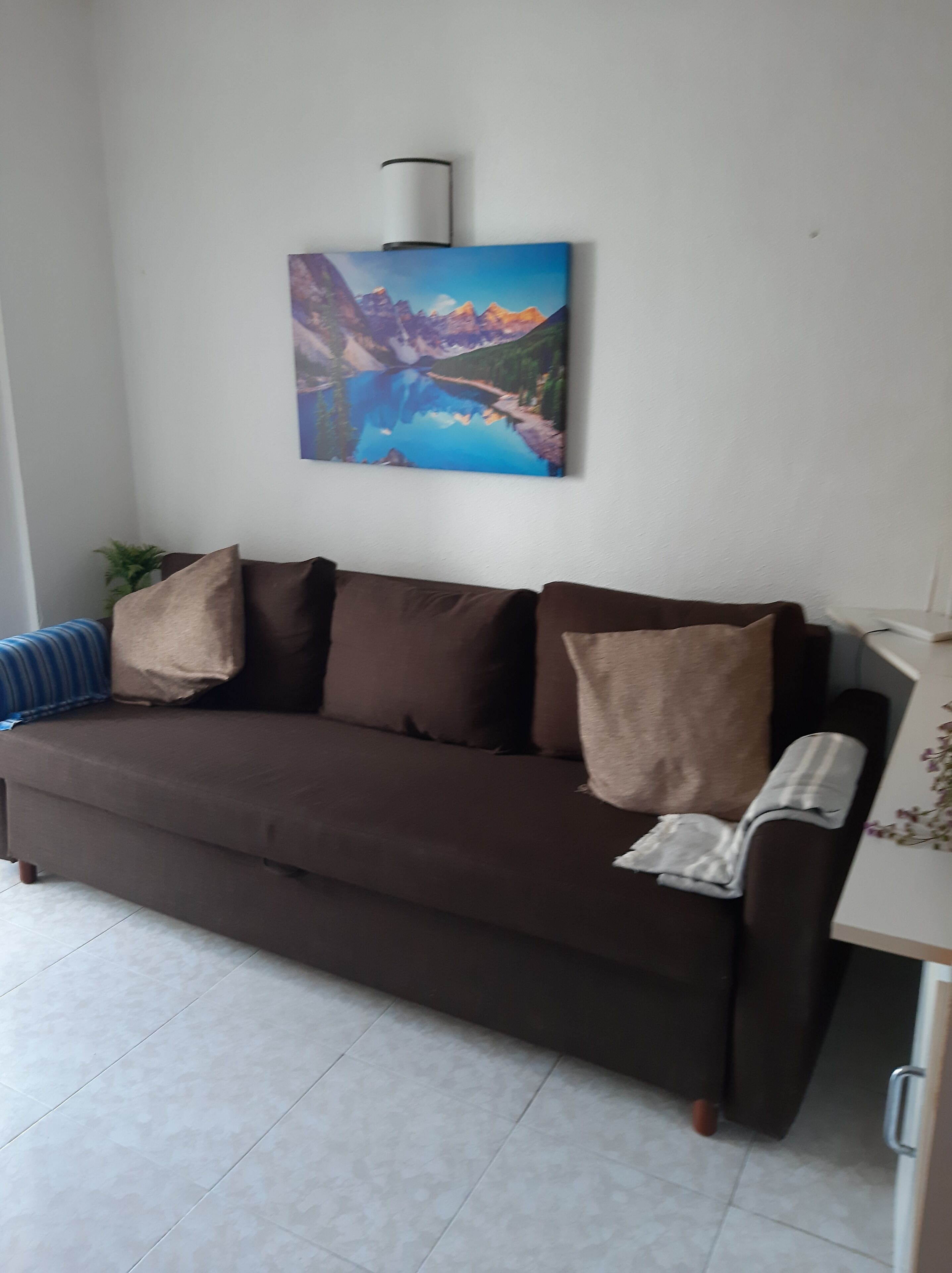 Um sofá marrom com várias almofadas, uma luminária na parede e uma pintura de uma paisagem de montanhas.