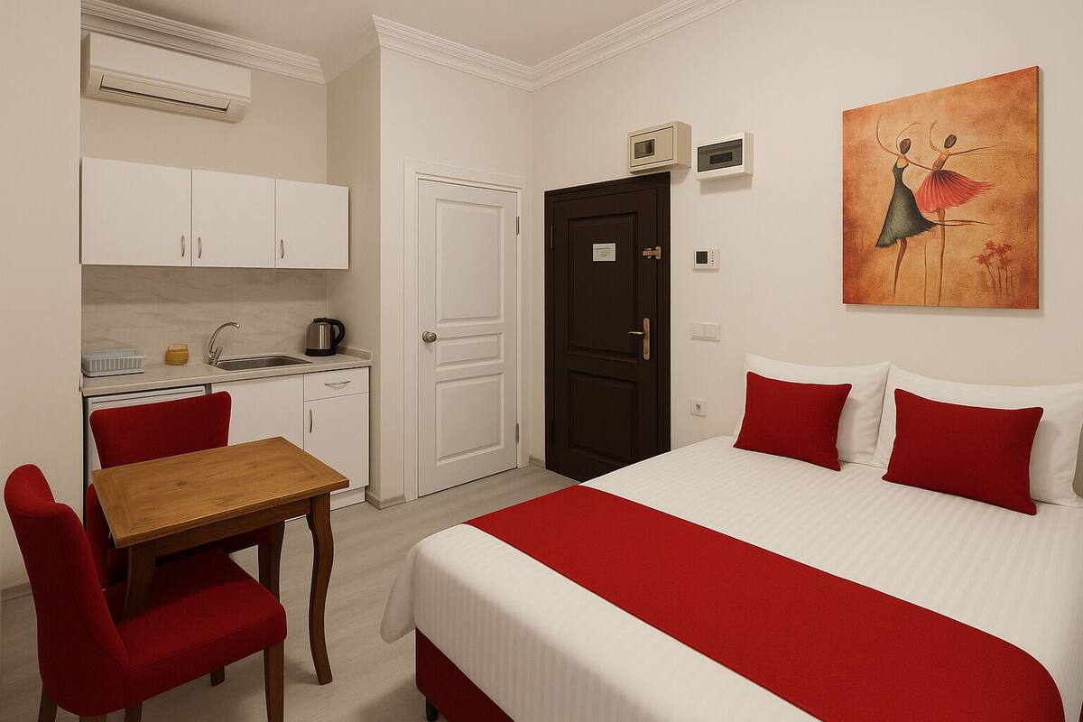 A hotel room with a bed, red pillows, a wooden table, red chairs, a kitchen area with cabinets, a microwave, and a painting on the wall.