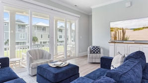 A spacious living room with a large TV, comfortable seating, and a view of a residential area through large sliding doors.