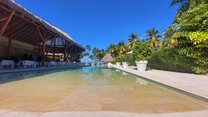 Una piscina cristalina con sillas de descanso y sombrillas, rodeada de palmeras y una estructura de techo de paja.