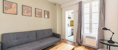 A living room with a grey sofa, wooden coffee table, and a lamp on a side table. There are framed pictures on the wall and a large window with curtains.