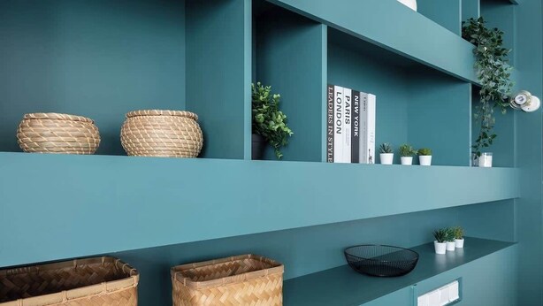 Uma parede cor de verde-água com prateleiras que exibem cestos trançados, livros e pequenas plantas em vasos.