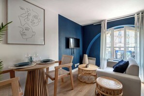 Una sala de estar moderna con una pared azul decorativa, un televisor de pantalla plana sobre la chimenea, una mesa de comedor redonda con sillas, y vista a un paisaje urbano a través de amplios ventanales.