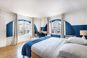 A hotel room with a bed, a desk, and a view of buildings through arched windows.