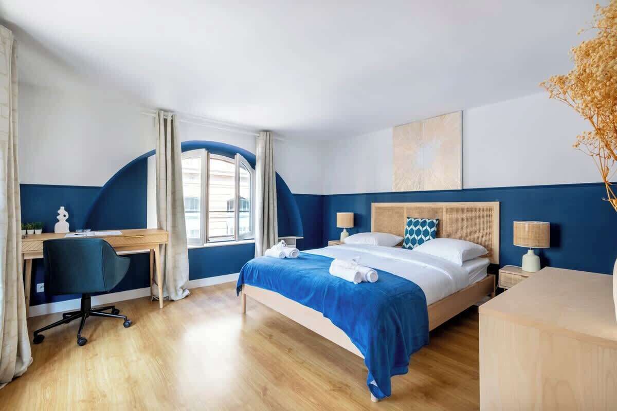 A hotel room with a bed, desk, and chair, a window with curtains, and a vase with dried flowers.