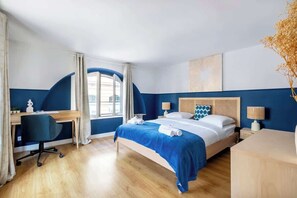 A hotel room with a bed, desk, and chair, a window with curtains, and a vase with dried flowers.