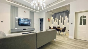 A modern living room with a sectional sofa, a flat-screen TV, a dining area with a table and chairs, and a hallway leading to a door. - 99 Mozilla Apartments (Abuja)