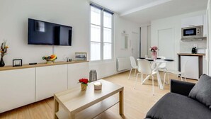 A modern living room with a flat-screen TV, a dining area with a table and chairs, and a kitchen area with a microwave.