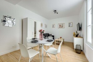 A modern dining area with a round table, white chairs, and a vase of flowers. There is a white cabinet and a television on a stand.