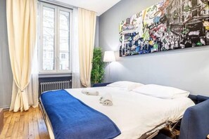 Habitación de hotel con cama, ventana con cortinas, radiador y un cuadro colgado en la pared.
