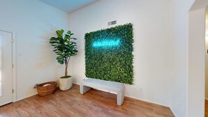 A room with a wooden floor, a bench, a potted plant, and a wall-mounted green wall with a neon sign.